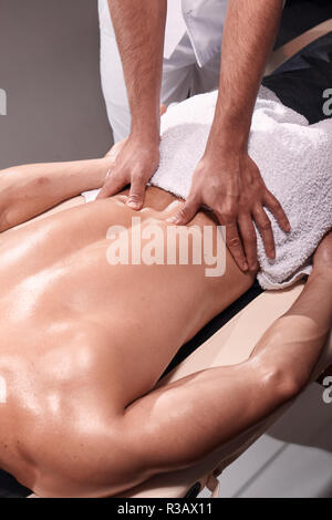 two young man, 20-29 years old, sports physiotherapy indoors in studio, photo shoot. Physiotherapist massaging patient lower back with his hands close Stock Photo