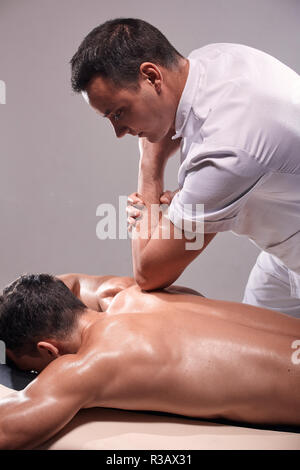 two young man, 20-29 years old, sports physiotherapy indoors in studio, photo shoot. Physiotherapist massaging muscular patient back with his elbow. Stock Photo
