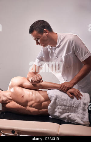 two young man, 20-29 years old, sports physiotherapy indoors in studio, photo shoot. Therapist masseur massaging arm of muscular patient laying on his Stock Photo