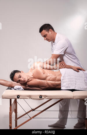 two young man, 20-29 years old, sports physiotherapy indoors in studio, photo shoot. Therapist masseur massaging arm of muscular patient laying on his Stock Photo