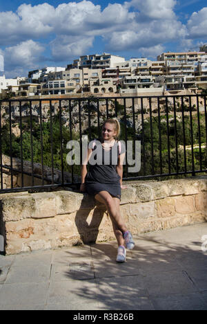 young blond girl in gray dress with pink backpack siting on the stone gate - teeneger or fashion concept Stock Photo