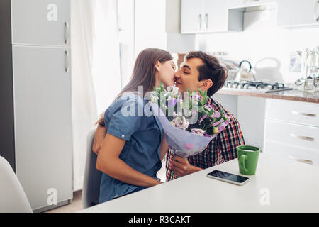 Young man gifting bouquet of flowers to his girlfriend in kitchen. Happy couple hugging. Romantic surprise at home Stock Photo