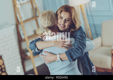 Long awaited meeting. Cheerful daughter hugging her adorable elderly mother standing with her back to camera while smiling and looking at you Stock Photo