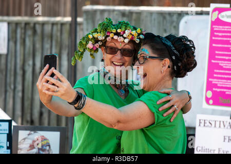 Two women embracing, happily smiling with both holding a cellphone, mobile phone, and talking selfie of themselves. One wears garland of hops. Stock Photo
