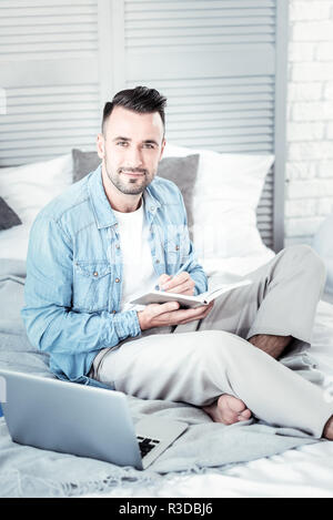 Watching you. Handsome bearded man keeping smile on his face and making notes while looking at camera Stock Photo