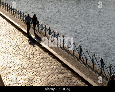 couple walking along a cobbled street next to a river with the sun behind Stock Photo