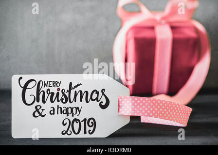 Label With English Calligraphy Merry Christmas And A Happy 2019. One Pink Gift With Ribbn. Cement Background. Stock Photo