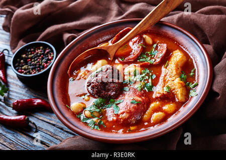 Fabada Asturiana Bean hearty Stew in an earthenware bowl with spoon on a rustic wooden table with brown cloth, chili peppers and parslaey, spanish cui Stock Photo