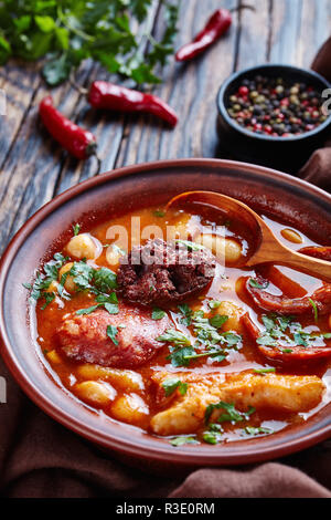 Fabada Asturiana Bean hearty Stew in an earthenware bowl with spoon on a rustic wooden table with brown cloth, chili peppers and parslaey, spanish cui Stock Photo