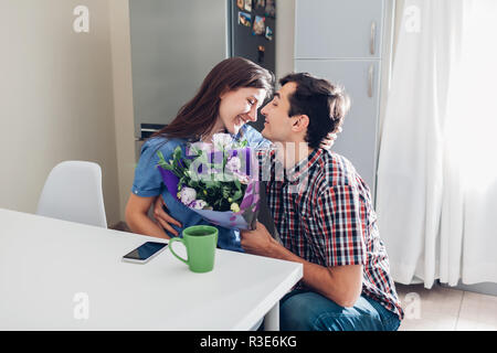 Young man gifting bouquet of flowers to his girlfriend in kitchen. Happy couple hugging. Romantic surprise at home Stock Photo