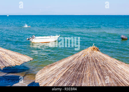 View on empty public beach with thatched sunshades, umbrellas and deckchairs along sandy coastline. Small jetty boat is anchored close to the beach. Stock Photo
