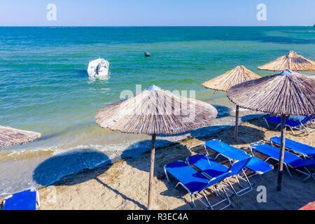 View on empty public beach with thatched sunshades, umbrellas and deckchairs along sandy coastline. Small jetty boat is anchored close to the beach. Stock Photo