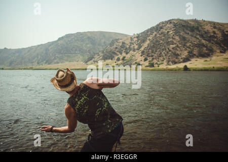 Candid photograph of man in a cowboy hat, skipping stones across the river as he takes a break from driving on his roadtrip to stretch his legs. Stock Photo