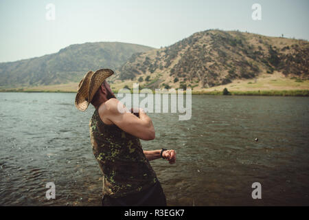 Candid photograph of man in a cowboy hat, skipping stones across the river as he takes a break from driving on his roadtrip to stretch his legs. Stock Photo