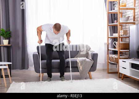 Disabled Young Man Using Crutches To Get Up From Sofa At Home Stock Photo