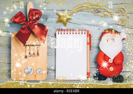 Fun background for the Christmas menu or recipes. Blank notebook, cutting board with utensils and miniature dishes and Santa Claus doll. Stock Photo