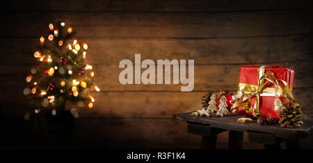 red gift box and christmas decoration on an old stool, blurry tree with lights in the background against a rustic wooden wall, copy space, panoramic f Stock Photo