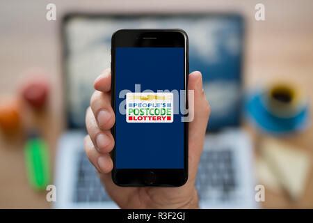 A man looks at his iPhone which displays the People’s Postcode Lottery logo, while sat at his computer desk (Editorial use only). Stock Photo