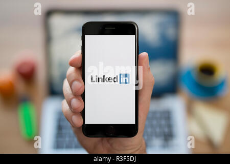 A man looks at his iPhone which displays the LinkedIn logo, while sat at his computer desk (Editorial use only). Stock Photo