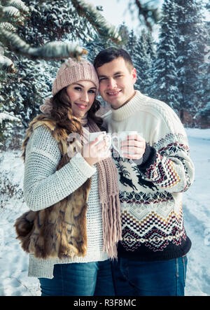 Couple hugs and holds cups of coffee with marshmallow in winter forest Stock Photo