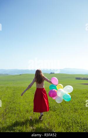 Happy girl in the meadows tuscan with colorful balloons, against the blue sky and green meadow. Tuscany, Italy Stock Photo