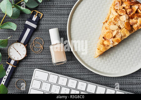 Cozy business composition with white computer keyboard, nail polish, rings, eucalyptus plant, women's watch and apple pie on plate over grey backgroun Stock Photo