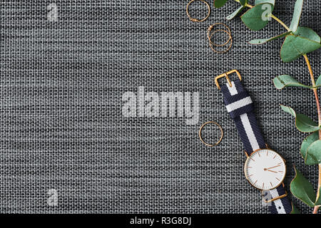 Women's watch with navy blue and white nylon strap, golden rings and eucalyptus on grey background. Top view with copy space for text.