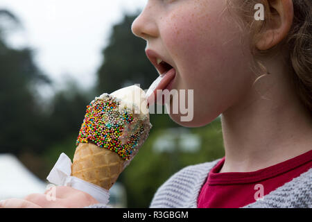 Six-year-old girl/child/kid licks an ice cream cone, decorated with hundreds and thousands, while it is melting rapidly.  (98) Stock Photo