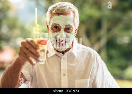 Funny senior man with clay facial mask holding a glass of juice. Man having spa facial treatment at retired age. Stock Photo
