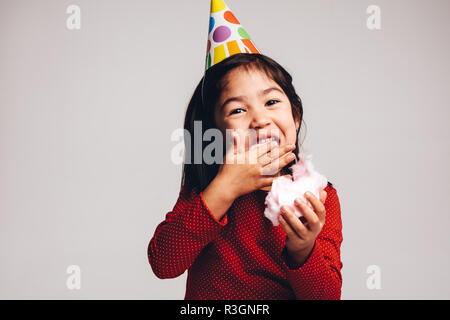 Portrait of a little girl eating sugar candy floss. Kid wearing a party cone cap enjoying cotton candy. Stock Photo