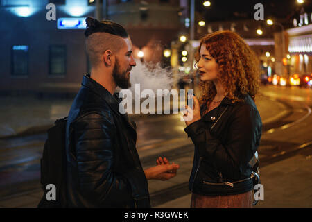 Couple in love on date. Boyfriend talking to girlfriend. She smokes an electronic cigarette and looks at him. They walk on streets of night city. Wet  Stock Photo