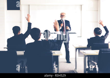 Business Audience raising hand up while businessman is speaking in training for Opinion with Meeting Leader in Conference Room Stock Photo
