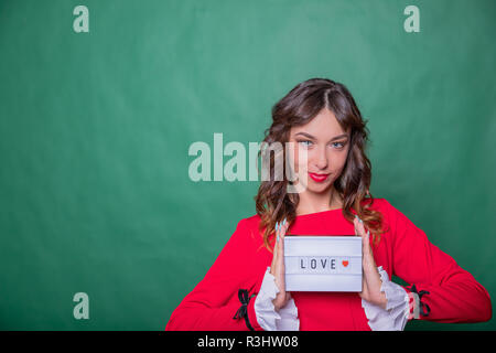 Advertising. Woman in red dress. Smiling woman showing board card with word love and red heart on green background. love text message.Positive human emotions face expressions.Happy valentines greeting concept Stock Photo