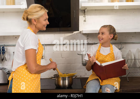 happy grandmother cooking spaghetti and looking at smiling granddaughter holding cookbook in kitchen Stock Photo