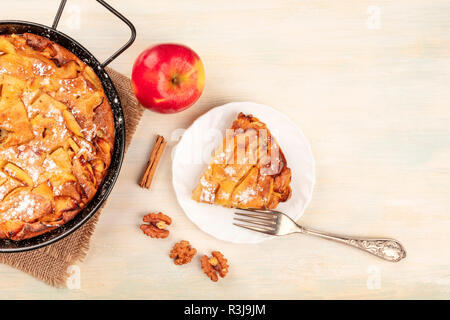 A photo of an apple pie in a pan and a slice of it on a plate, shot from the top on a rustic wooden background with an apple, a cinnamon stick, walnut Stock Photo