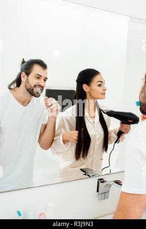 handsome adult man brushing teeth while wife drying hair with hairdryer Stock Photo