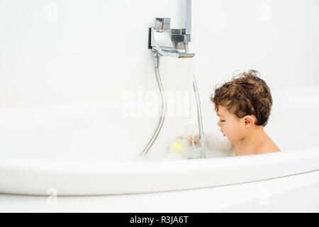 Toddler boy sitting in white bathroom and playing with bath toys Stock Photo