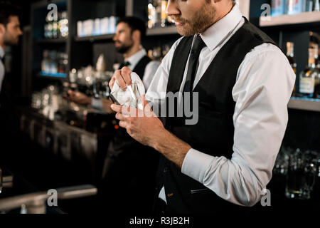 Old pub with bartender cleaning the glass with white towel on background.  Empty name tag badge on the shirt uniform. Barman at his working place.  Stock Photo