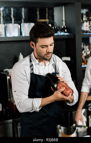 handsome barman in apron preparing cocktail in glass with shaker Stock Photo