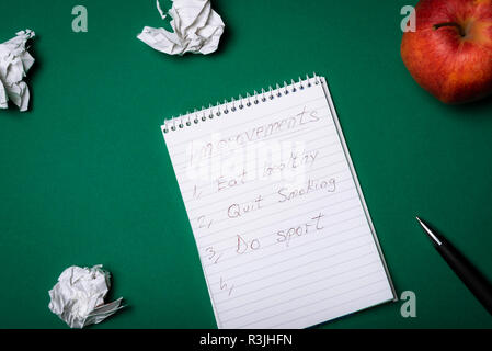 Spiral notebook with a list of improvements, eat healthy, quit smoking, do sport, and crumpled pages and an apple. Top view. Resolutions. Stock Photo