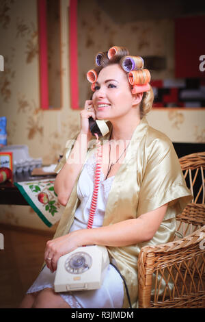 Cute woman in bathrobe and hair curlers talking on the phone while sitting in the kitchen. Retro and vintage. Stock Photo