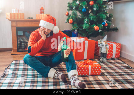 Bored and drunk young man sit on blanket on floor and look at bottle. He holds one hand under chin. He is falling asleep Stock Photo