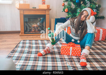 Young woman sits on floor and sleeps. She holds green bottle of alcohol in hand. There is a box with present between her legs. She is drunk Stock Photo