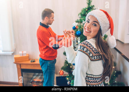 Nice picture of attractive young woman standing and looking on camera. She smiles. Guy decorates Christmas tree. He is concentrated Stock Photo