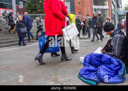 Manchester, UK. 23 November, 2018. Black Friday Sales Weekend.  Homeless man; City centre holiday shopping season, retail shops, stores, Christmas shoppers, discount sale shopping, female consumer spending on Black Friday weekend considered to be the biggest shopping event of the year.   U.K. retailers have embraced the USA style sale bonanza, with many customers left surprised by bargain discounts, enjoying a Xmas spend & carrying a number bags, gifts, presents and sale items. Credit: MediaWorldImages/Ģ����ýLiveNews. Stock Photo
