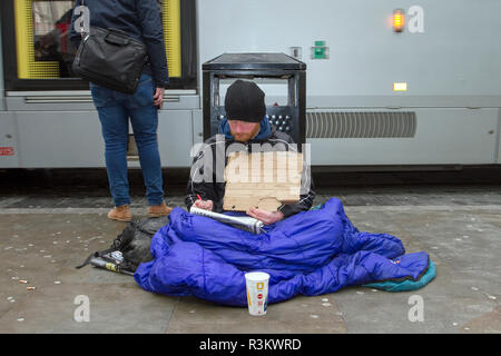 Manchester, UK. 23 November, 2018. Black Friday Sales Weekend.  Homeless man; City centre holiday shopping season, retail shops, stores, Christmas shoppers, discount sale shopping, female consumer spending on Black Friday weekend considered to be the biggest shopping event of the year.   U.K. retailers have embraced the USA style sale bonanza, with many customers left surprised by bargain discounts, enjoying a Xmas spend & carrying a number bags, gifts, presents and sale items. Credit: MediaWorldImages/Ģ����ýLiveNews. Stock Photo