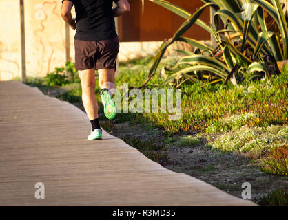 A man runs on a boardwalk with his right feet facing up. Brown shorts, green sneakers. Rear view. Stock Photo