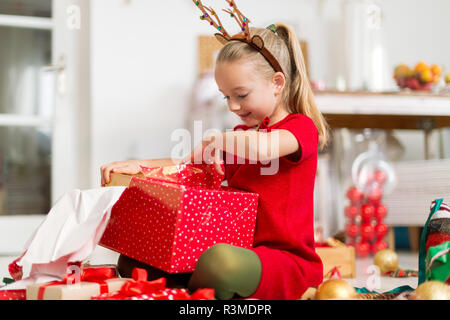 Cute super excited young girl opening large red christmas present while sitting on living room floor. Candid family christmas time lifestyle backgroun Stock Photo