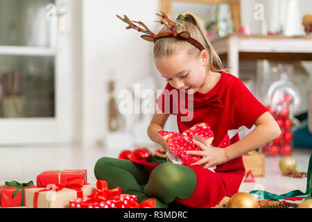 Cute super excited young girl opening large red christmas present while sitting on living room floor. Candid family christmas time lifestyle backgroun Stock Photo