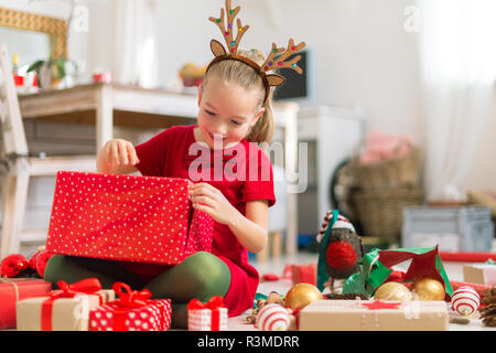 Cute super excited young girl opening large red christmas present while sitting on living room floor. Candid family christmas time lifestyle backgroun Stock Photo